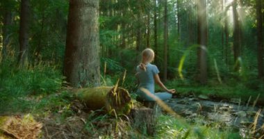 Sessiz bir ormanda akıntıya karşı meditasyon yapan bir kadın. Güneş ışığı yavaşça uzun ağaçların arasından süzülüyor..