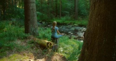 Kadın yoga pozunda oturuyor ormandaki bir ağaç kütüğünde akarsuyun yanında yemyeşil ve uzun ağaçlarla çevrili..