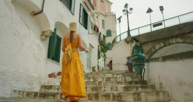 Beyaz binaların ve büyük boyalı vazoların yanından geçen yıpranmış taş merdivenlerde yürüyen bir kadın..