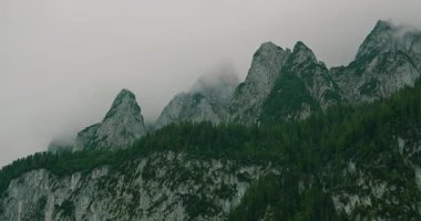 Aşağıda bulutlu bir gökyüzünün altında yeşil ormanlarla kaplı dramatik dağ zirveleri..