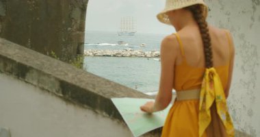 Elinde harita olan kadın, Atrani şehrinin kayalık kıyılarındaki yelkenli gemi ve teknelere bakıyor..