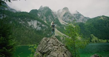 Kadın kolları havada kayalık bir tepede duruyor, yüksek dağlara bakıyor ve Gosau Gölü 'nü çevreleyen yemyeşil yeşilliklere bakıyor..