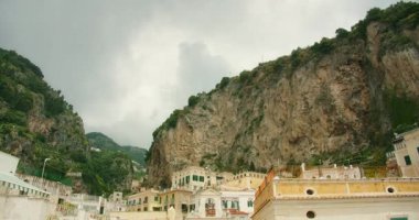 Atrani 'deki eski kasaba binalarının tepesinde bulutlu gökyüzünün altında yeşil ağaçlarla dolu bir uçurum..