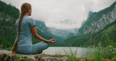 Huzurlu bir gölde meditasyon yapan bir kadın, görkemli dağlar ve bulutlarla çevrili, derin bir yansımaya dalmış..