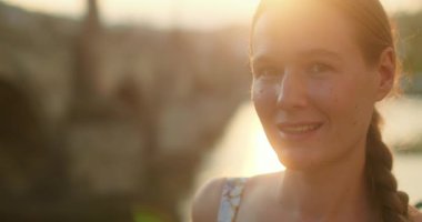 Charles Köprüsü 'nün yanında gün batımını yansıtan nehirle birlikte duran, sıcak altın güneş ışığında gülümseyen kadın..