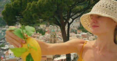 Arka planda Amalfi kasabası ve kıyı şeridi olan limon tutan kadının yakın çekimi.