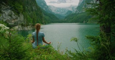 Kadın çimenli bir göl kıyısında oturuyor, Avusturya 'daki Gosau Gölü' nde dağ manzarasıyla meditasyon yapıyor..