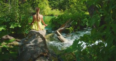 Kadın, yemyeşil ormanlarla çevrili bir derenin üzerindeki kütük üzerinde meditasyon yapıyor..