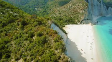 Kefalonia adasında, yemyeşil kayaların ve ziyaretçilerin dinlendiği uzak beyaz bir plajın üzerindeki yüksek bir uçurumda duran bir kadın..