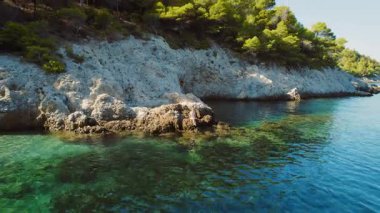 Açık yeşil deniz ve çam ağaçlarıyla kaplı uçurumların yanındaki kayalıklarda oturan kadın..