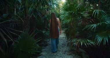 Çok uzun saçlı bir kadın palmiye ağacı ormanında, gür bir orman yolunda yürüyor..