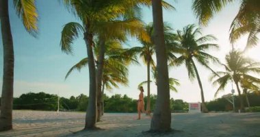 Gün batımında voleybol ağının ve palmiye ağaçlarının yanından geçen bir kadın..