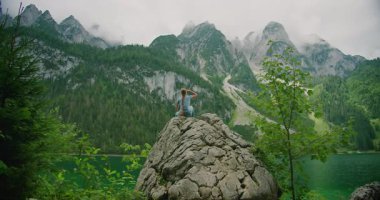 Kadın, zümrüt göle bakan engebeli bir kayaya tünemiş. Gosausee, Avusturya 'daki bulutlu tepelere bakıyor..