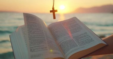 Açık kitap ve haç kolye sıcak yaz havasında gün batımında deniz kenarında tutulur..