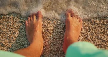 Gün batımında kıyıya vuran dalgalar gibi çıplak ayakla çakıllı sahilde duran bir insan..