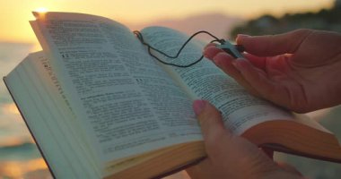 Alacakaranlıkta sahilde elinde haç kolyeyle İncil okuyan birinin yakın çekimi..