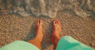 Gün batımında deniz dalgaları yaklaşırken sakin çakıl taşlı sahilde duran yalın ayaklı adam..