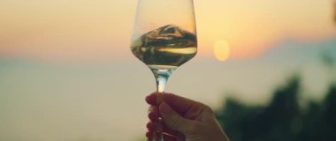 Gün batımına karşı yükselen beyaz şarap kadehi dağlar ve sahil manzaralı..