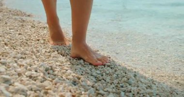 Hırvatistan 'da yaz aylarında sakin deniz dalgalarıyla çakıllı sahilde çıplak ayaklı bir kadın yürüyor.