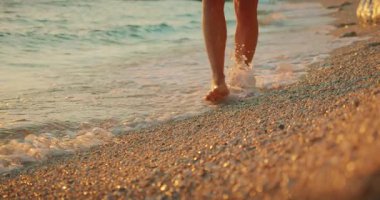 Altın gün batımında, çakıllı kıyı şeridi boyunca yalınayak yürüyen bir kadın..