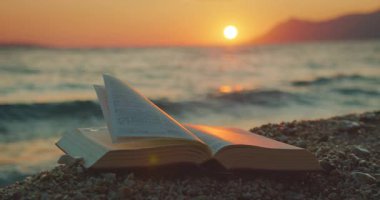 Gün batımında arka planda okyanus dalgaları olan çakıllı bir plajda açık bir kitap dinleniyor..