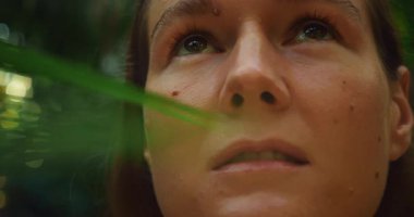 Yeşil bir tropikal ormandaki yaprakların arasından bakan bir kadın yüzü..