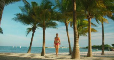 Kadın güvenle yürüyor kumlu bir sahil boyunca Palmiye ağaçları ve deniz manzarasıyla kaplı.