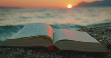 Gün batımında çakıl taşlı bir sahilde, arka planda deniz dalgaları olan kutsal kitap..
