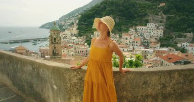 Kadın Amalfi limanına bakan terasta poz veriyor arkasında renkli bir kasaba var..