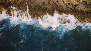 Kaba deniz dalgalarının havadan görünüşü altın kahverengi kayalık bir kıyıya çarpıyor..