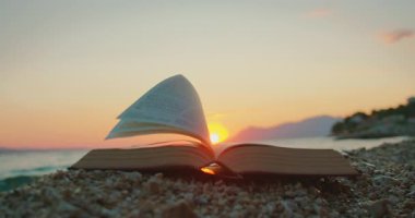 Kitap sayfaları kayalık sahilde alacakaranlıkta güneş dağların ardında batarken çırpınır..