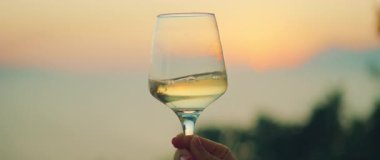 Elinde pembe günbatımı gökyüzü ve yumuşak arka planda tepeler olan beyaz şarap kadehi tutan kişi..