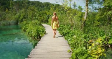 Yazın Plitvice Parkı 'nda, turkuaz gölün yanında, tahta patika boyunca tek başına yürüyen bir kadın..