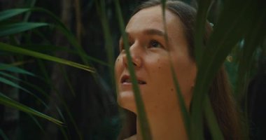 Bir kadın yemyeşil orman ortamında tropikal yaprakların arasından yukarı bakıyor..