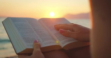 Gün batımında kumsalda İncil okuyan bir kadın. Parmakları sayfada..