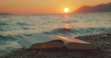 Hırvatistan 'da yaz aylarında deniz üzerinde gün batımı olan çakıllı bir plajda uzanan açık bir kitap.