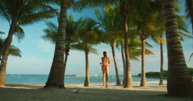 Mavi okyanusa doğru palmiye ağaçlarıyla kaplı beyaz kumsalda koşan bir kadın..
