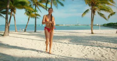 Bikinili gülümseyen kadın, turkuaz suyun altında, palmiye ağaçlarının altında, beyaz kumsalda yürüyor..