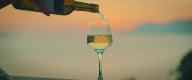 Gün batımında, arka planda deniz manzarası olan beyaz şarap bardağa dökülüyor..