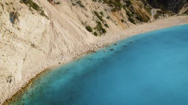 Dik uçurumların altında parlak turkuaz deniz suyuyla uzun bir sahil manzarası..