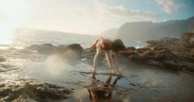 Gün batımında kayalık sahilde akıntı kenarında yansıyarak dans eden bir kadın..