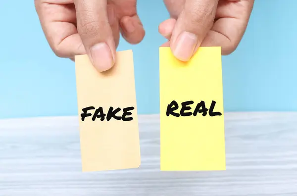 stock image Hand holding the words Fake or Real on yellow shaped paper isolated on blue background. Real or fake business concept