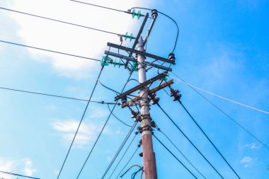 Yüksek voltajlı elektrik direği mavi gökyüzü arka planına sahip yerleşim bölgesinde birçok kablo var. Direğin üzerindeki kablolar oldukça düzgün..