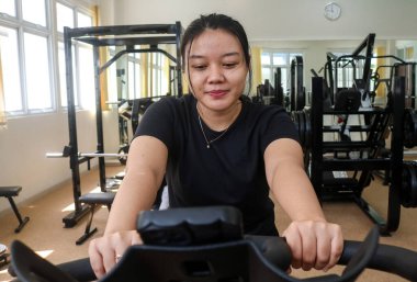 Spor kıyafetli gülümseyen genç bir kadının spor salonunda sabit bir bisiklete binişini ön planda görmek. Yeni başlayanlar için çalış
