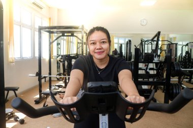 Spor kıyafetli gülümseyen genç bir kadının spor salonunda sabit bir bisiklete binişini ön planda görmek. Yeni başlayanlar için çalış