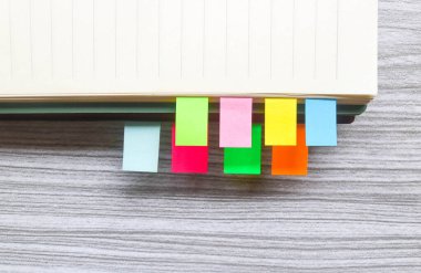 Kitap yığınları arasında renkli yapışkan notlar. Not defterine renkli yapışkan not yer imleri iliştirilmiş. Tahta masadaki kitap ayracları