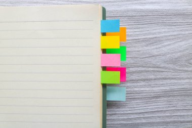 Kitap yığınları arasında renkli yapışkan notlar. Not defterine renkli yapışkan not yer imleri iliştirilmiş. Tahta masadaki kitap ayracları