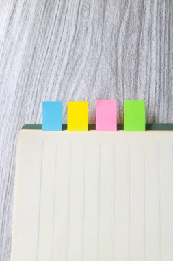 Kitaplar arasında renkli yapışkan notlar. Not defterine renkli yapışkan not yer imleri iliştirilmiş. Tahta masadaki bir kitabın yer imleri