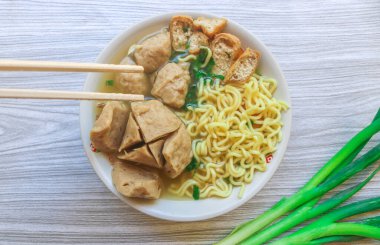 Indonesian culinary soup meatballs filled with noodles, tofu, celery and egg served in a white bowl on a gray wooden table. Close up of meatballs in sauce in a bowl with chopsticks and spoon clipart