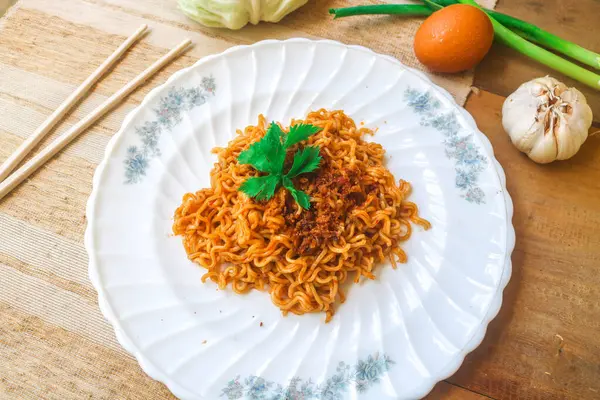 stock image Top view of spicy fried instant noodles topped with fried onions and vegetables in a white plate on a wooden table. Spicy variant of fast food noodles